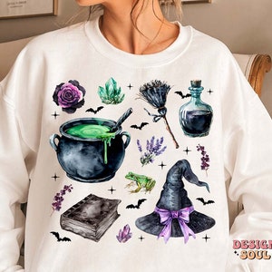 May include: White sweatshirt with a watercolor design of a witch's cauldron, a broom, a witch's hat, a bottle of potion, crystals, flowers, and bats. The design is in shades of green, purple, black, and white.