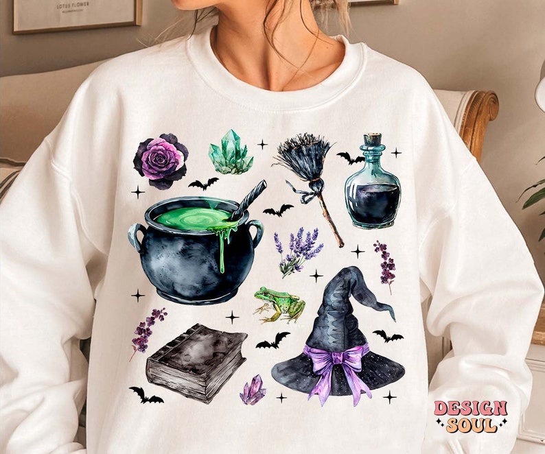 May include: White sweatshirt with a watercolor design of a witch's cauldron, a broom, a witch's hat, a bottle of potion, crystals, flowers, and bats. The design is in shades of green, purple, black, and white.