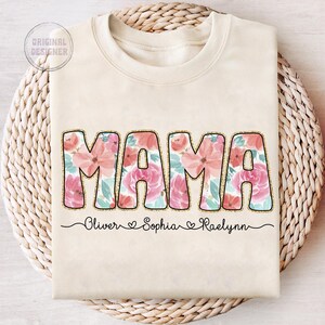 May include: A beige sweatshirt with the word "MAMA" in large, pink floral letters. The names "Oliver", "Sophia", and "Raelynn" are written below the word "MAMA" in a cursive font.