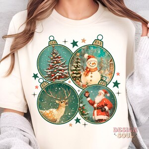 May include: A white t-shirt with a Christmas design featuring four baubles. Each bauble has a different Christmas scene: a Christmas tree, a snowman, a reindeer, and Santa Claus. The baubles are surrounded by stars and snowflakes.