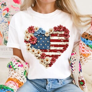 May include: White t-shirt featuring a heart-shaped design composed of red, white, and blue flowers and the American flag. The flag design has a distressed, vintage look. The shirt is a classic crew neck style.