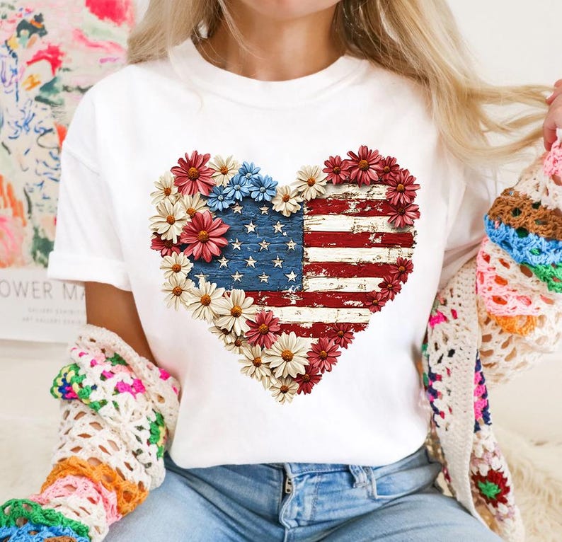May include: White t-shirt featuring a heart-shaped design composed of red, white, and blue flowers and the American flag. The flag design has a distressed, vintage look. The shirt is a classic crew neck style.