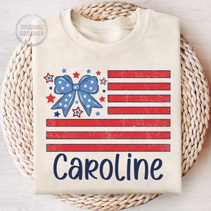 May include: A beige t-shirt with a distressed American flag design featuring a blue bow with white stars. The name "Caroline" is printed below the flag in blue.