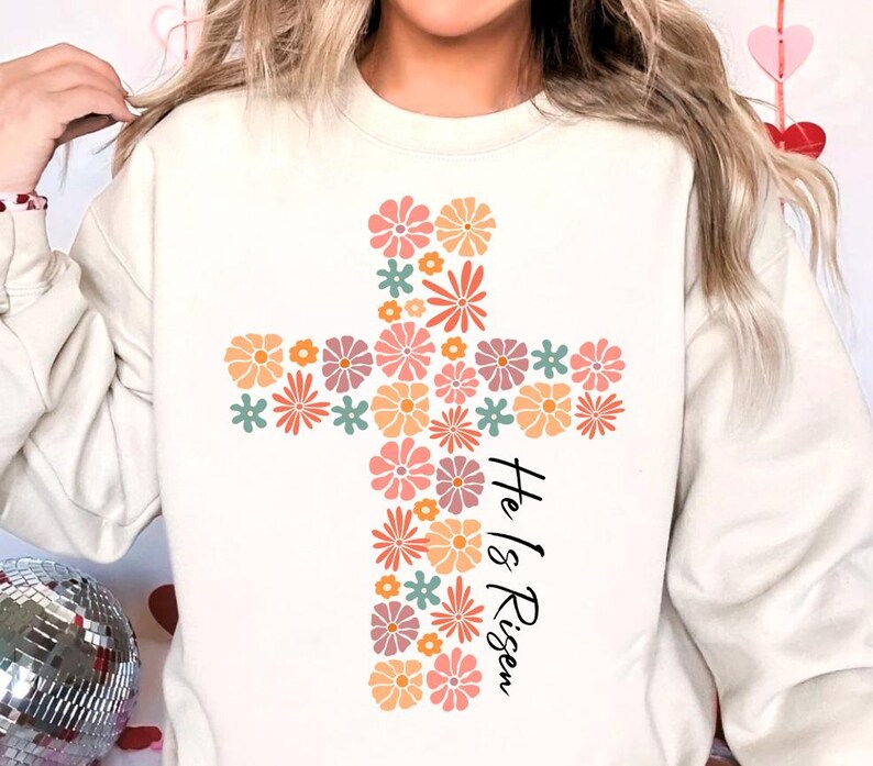 May include: White sweatshirt with a floral cross design and the text "He is Risen" in black.