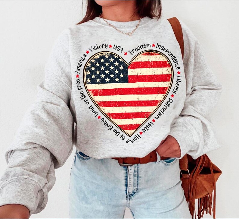 May include: Gray sweatshirt with a red, white, and blue heart-shaped American flag design. The heart is surrounded by a circle of text that reads "Home of the Brave Land of the Free America Victory USA Freedom Independence Liberty Patriotism Unity".