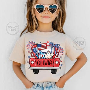 May include: A beige t-shirt featuring a red truck graphic with patriotic stars, an American flag, and fireworks. The name "OLIVIA" is printed on the truck. The shirt is worn by a young person wearing heart-shaped sunglasses.