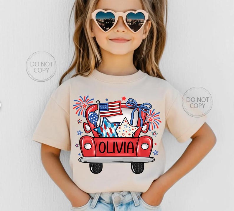 May include: A beige t-shirt featuring a red truck graphic with patriotic stars, an American flag, and fireworks. The name "OLIVIA" is printed on the truck. The shirt is worn by a young person wearing heart-shaped sunglasses.