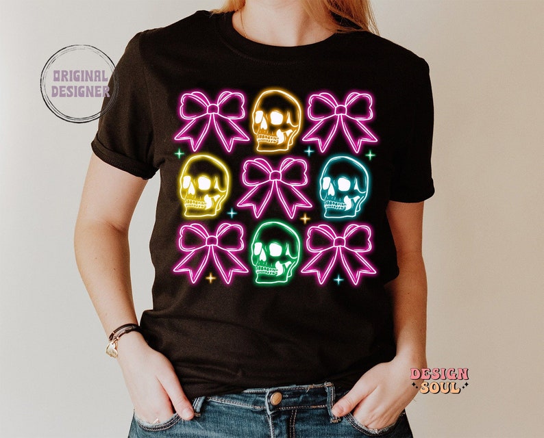 May include: Black t-shirt with a neon design of skulls and bows. The skulls are yellow, green, and blue, and the bows are pink, purple, and teal.