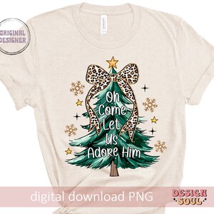 May include: A beige t-shirt with a green Christmas tree design. The tree is decorated with a leopard print bow and the words "Oh Come Let Us Adore Him".