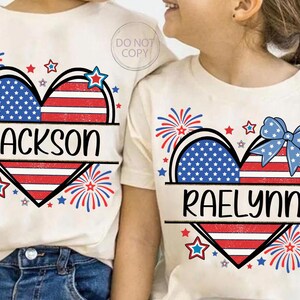 May include: Off-white t-shirts with patriotic heart designs. One shirt features the name "Jackson" and the other "Raelynn". The hearts are split into sections of blue stars and red and white stripes, with fireworks and star accents.