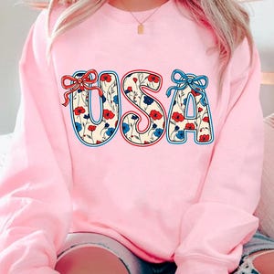 May include: Pink sweatshirt with a floral patterned "USA" graphic. The letters are outlined in red and blue, and the flowers are red, blue, and white.