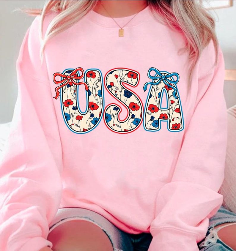 May include: Pink sweatshirt with a floral patterned "USA" graphic. The letters are outlined in red and blue, and the flowers are red, blue, and white.
