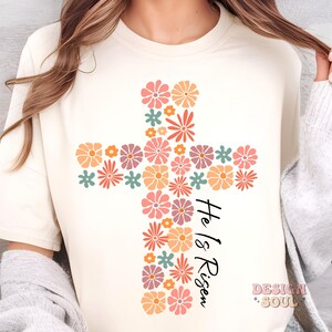 May include: A white t-shirt with a floral cross design and the text "He is Risen" in black lettering.