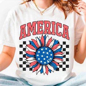 May include: White t-shirt with a red, white, and blue sunflower design. The sunflower has a blue center with white stars and is surrounded by red, white, and blue petals. The word "AMERICA" is printed in red above the sunflower.
