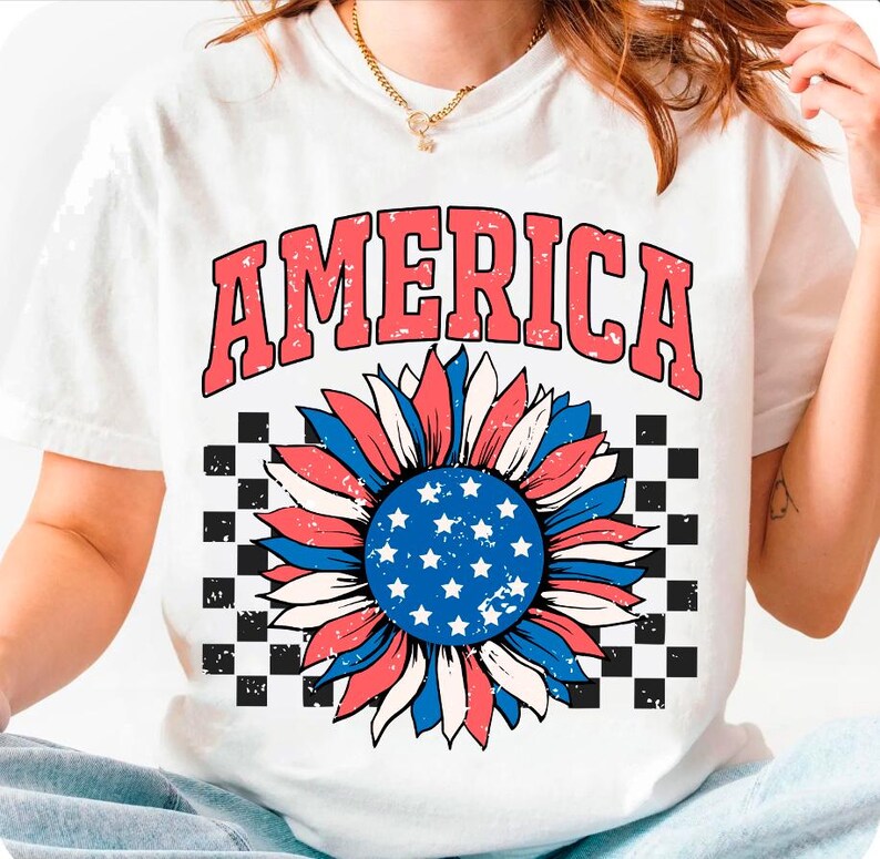 May include: White t-shirt with a red, white, and blue sunflower design. The sunflower has a blue center with white stars and is surrounded by red, white, and blue petals. The word "AMERICA" is printed in red above the sunflower.