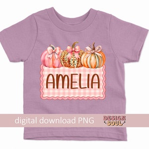 May include: A purple t-shirt with a pink and orange pumpkin design and the name Amelia in brown text on a pink and white checkered background.