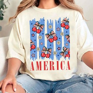 May include: A white t-shirt with a blue and red graphic design featuring cherries, stars, and daisies. The graphic design is reminiscent of the American flag. The text "AMERICA" is printed in red below the graphic design.