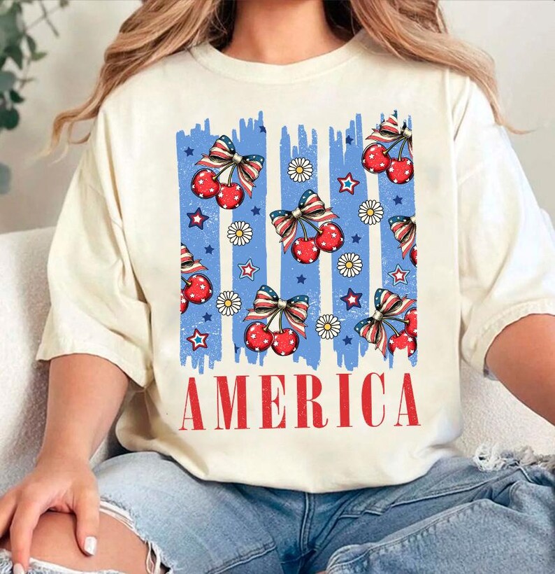 May include: A white t-shirt with a blue and red graphic design featuring cherries, stars, and daisies. The graphic design is reminiscent of the American flag. The text "AMERICA" is printed in red below the graphic design.