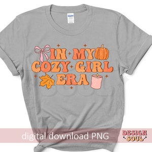 Puede incluir: Camiseta gris con el texto "In My Cozy Girl Era" en letras naranja y rosa. El dise&ntilde;o tambi&eacute;n incluye una calabaza, una vela y una hoja.
