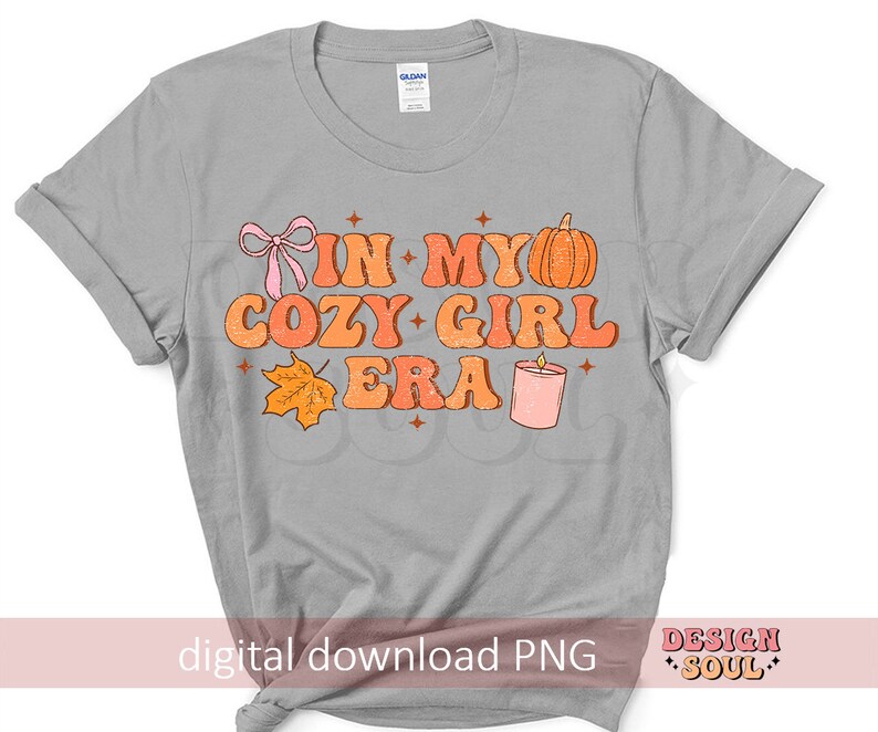 Puede incluir: Camiseta gris con el texto "In My Cozy Girl Era" en letras naranja y rosa. El dise&ntilde;o tambi&eacute;n incluye una calabaza, una vela y una hoja.