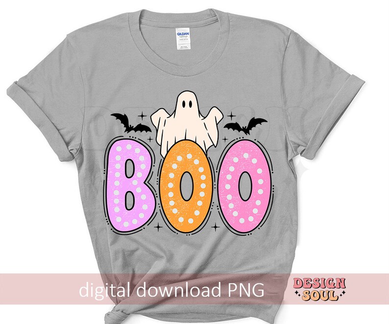 Op de afbeelding: Een grijs T-shirt met een spook en het woord "BOO" in roze en oranje stippen. De letters zijn zwart omrand. Boven het woord "BOO" bevinden zich twee zwarte vleermuizen.