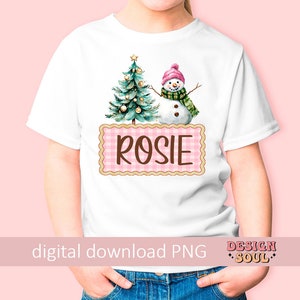 May include: A white t-shirt with a pink and white checkered box that says "Rosie" in brown text. The box is decorated with a watercolor illustration of a Christmas tree and a snowman.