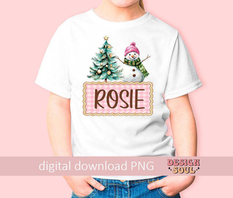 May include: A white t-shirt with a pink and white checkered box that says "Rosie" in brown text. The box is decorated with a watercolor illustration of a Christmas tree and a snowman.