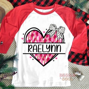 May include: A white and red baseball tee shirt with a pink and gold heart design. The heart has a black and white polka dot bow on top and the name "Raelynn" written across the center.