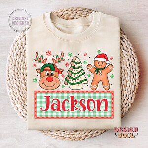 May include: A beige sweatshirt with a Christmas-themed design featuring a reindeer, a gingerbread man, and a Christmas tree. The design includes the name "Jackson" in red letters on a green and white checkered background.