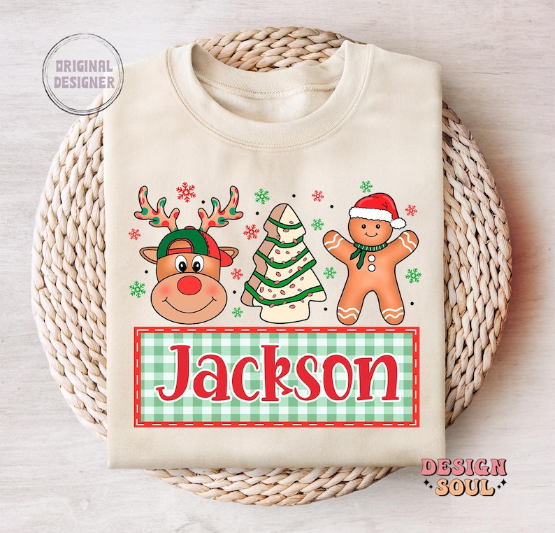 May include: A beige sweatshirt with a Christmas-themed design featuring a reindeer, a gingerbread man, and a Christmas tree. The design includes the name "Jackson" in red letters on a green and white checkered background.