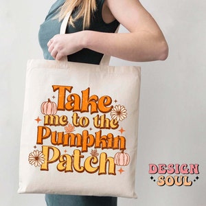 May include: A white tote bag with the text "Take me to the Pumpkin Patch" in orange and yellow letters. The text is surrounded by small orange pumpkins and flowers. The bag is being held by a person.
