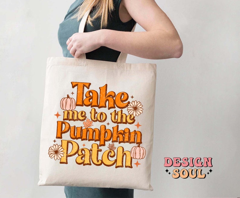 May include: A white tote bag with the text "Take me to the Pumpkin Patch" in orange and yellow letters. The text is surrounded by small orange pumpkins and flowers. The bag is being held by a person.