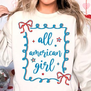 Peut inclure: Sweat-shirt blanc avec un motif graphique bleu et rouge qui dit "all american girl" avec des &eacute;toiles et des n&oelig;uds.
