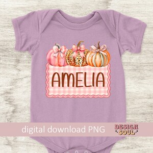 May include: A purple baby onesie with a pink and white checkered design and three pumpkins. The pumpkins are decorated with pink bows and have a leopard print, orange, and pink color scheme. The text "Amelia" is written in brown letters on the checkered design.