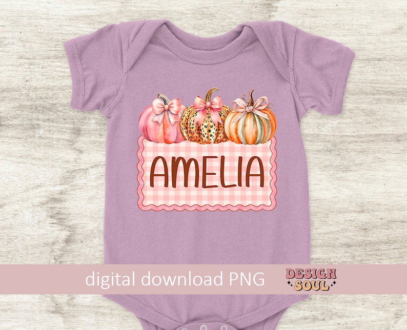 May include: A purple baby onesie with a pink and white checkered design and three pumpkins. The pumpkins are decorated with pink bows and have a leopard print, orange, and pink color scheme. The text "Amelia" is written in brown letters on the checkered design.