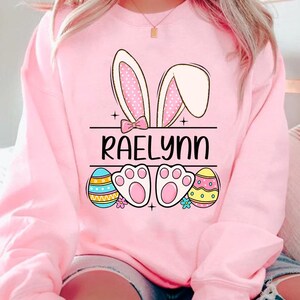 May include: Pink sweatshirt with a bunny ear design, Easter eggs, paw prints, and the name "RAELynn" printed on the front.