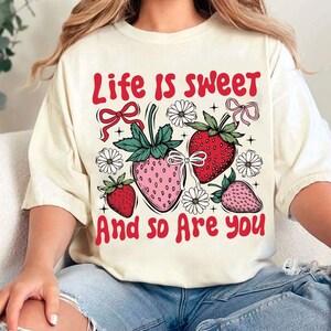 May include: A white t-shirt with a red graphic that says "Life is sweet and so are you". The graphic also features strawberries and flowers.