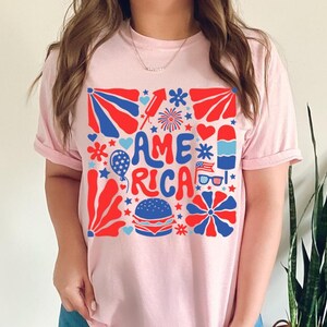 以下が含まれることがあります： レトロなスタイルで「アメリカ」という言葉が書かれたピンクのTシャツ。赤、白、青の星、花火、その他の愛国的なイメージに囲まれています。
