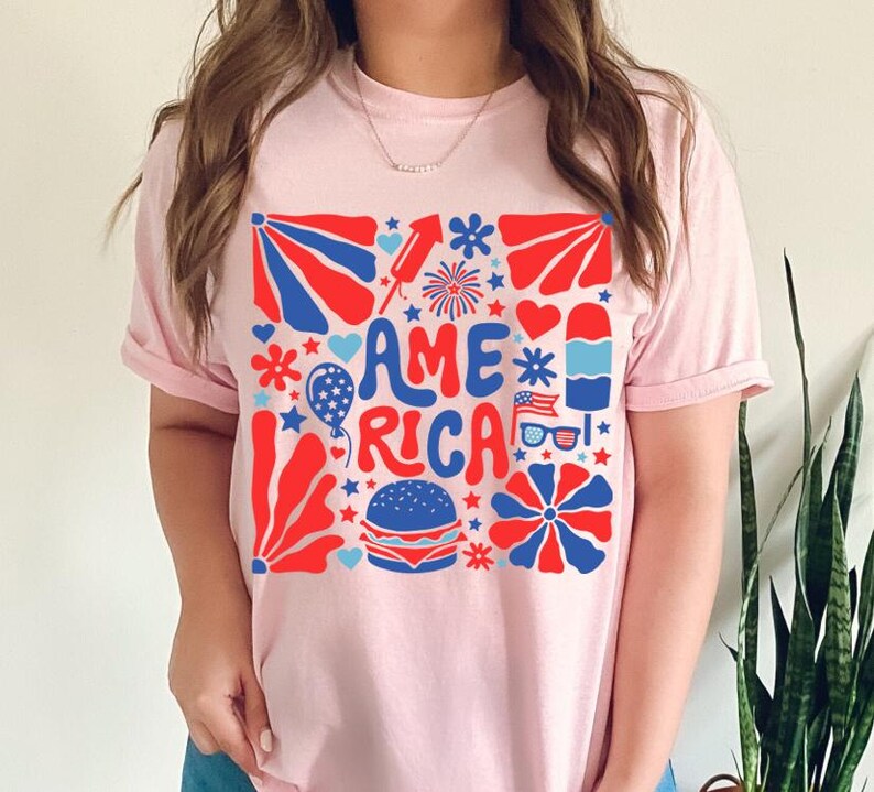 以下が含まれることがあります： レトロなスタイルで「アメリカ」という言葉が書かれたピンクのTシャツ。赤、白、青の星、花火、その他の愛国的なイメージに囲まれています。