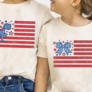 May include: Two white t-shirts with a red, white, and blue American flag design. One shirt features a blue dinosaur with sunglasses and the other shirt features a blue bow.