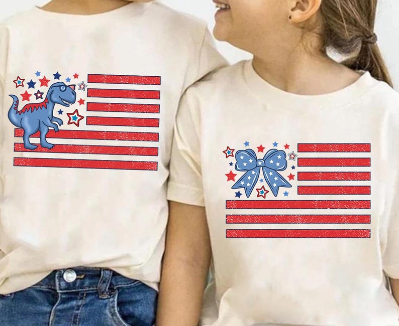 May include: Two white t-shirts with a red, white, and blue American flag design. One shirt features a blue dinosaur with sunglasses and the other shirt features a blue bow.