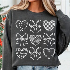 May include: A charcoal gray sweatshirt with a white design of nine hearts and bows. The hearts are filled with different patterns, including polka dots, checkered, and stripes.