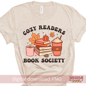 May include: A beige t-shirt with the text "Cozy Readers Book Society" in brown lettering. The design features a stack of books, a pumpkin, a candle, and a cup of coffee with whipped cream.