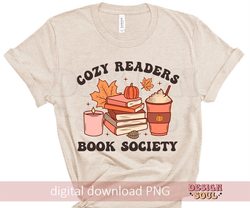 May include: A beige t-shirt with the text "Cozy Readers Book Society" in brown lettering. The design features a stack of books, a pumpkin, a candle, and a cup of coffee with whipped cream.