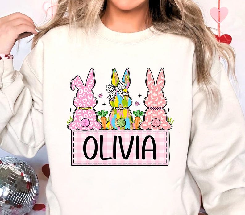 May include: White sweatshirt with a pink, blue, and green bunny rabbit design. The bunnies are sitting on a pink and white checkered background with the name "Olivia" written in black.