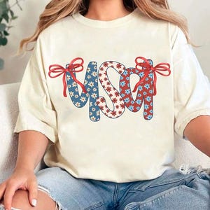 May include: A white t-shirt with a red, white, and blue USA graphic. The letters are made of a floral pattern with red and blue flowers. The graphic has red bows on each side.