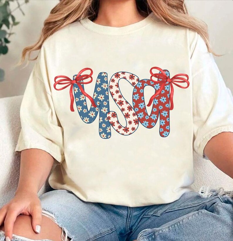 May include: A white t-shirt with a red, white, and blue USA graphic. The letters are made of a floral pattern with red and blue flowers. The graphic has red bows on each side.