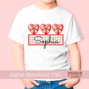 May include: White t-shirt with three red hearts with bows and a red and white gingham banner with the name "Sophia" printed on it.
