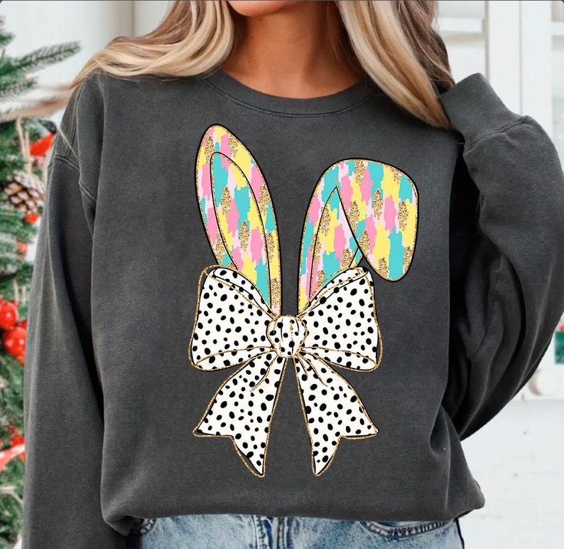 May include: A charcoal gray sweatshirt with a colorful bunny ear design and a black and white polka dot bow. The bow is tied around the bunny ears.