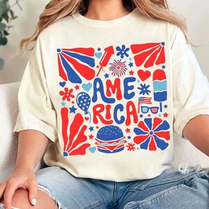 以下が含まれることがあります： 赤、白、青のグラフィックデザインが施された白いTシャツ。グラフィックにはレトロなフォントで「AMERICA」という言葉が書かれており、星、ハート、花火、その他の愛国的なイメージで囲まれています。